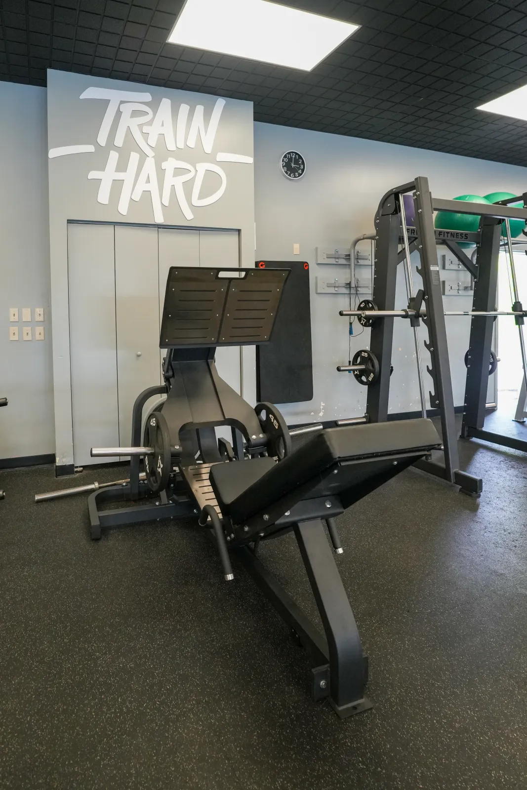 Plate-loaded leg press and Smith machine below the Train Hard mural at BR Fitness in East Anchorage